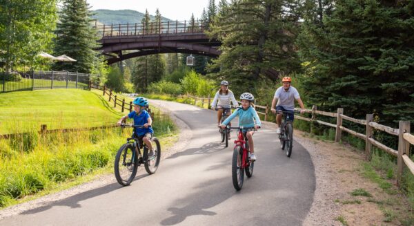 An adult and two children in helmets ride mountain bikes down a paved path in Vail. A brown footbridge is seen behind them.