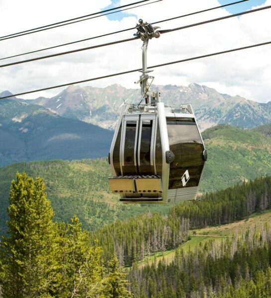 A silver gondola with a Vail logo on it heads up the mountain on a series of cables. Green trees and foliage blanket the terrain in the background.