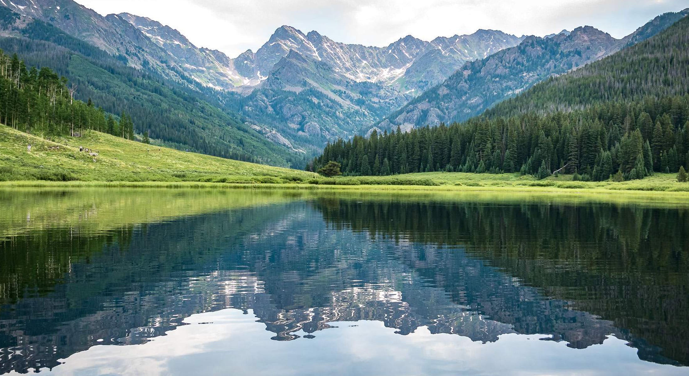 Rugged mountains above are reflected in Piney Lake, near Vail, Colorado. Green grass and pine trees cover the landscape.