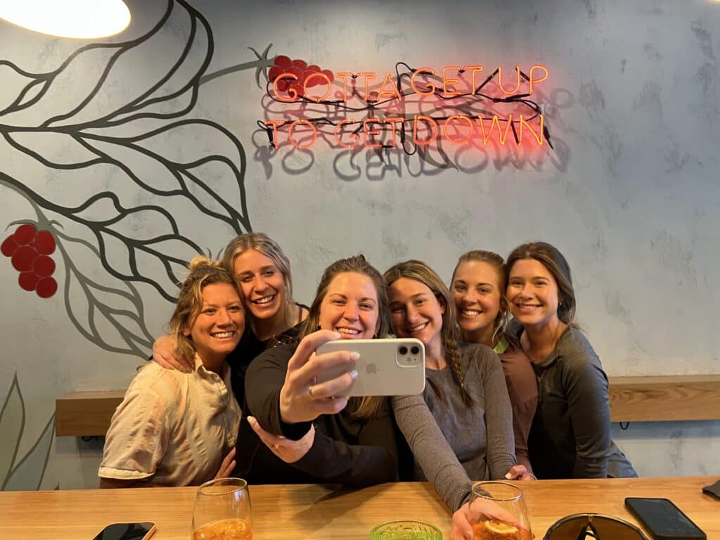 A group of 6 women standing in front of a bar counter take a selfie in front of a neon sign reading "gotta get up to get down."