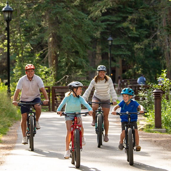 A family riding rental bikes down a tree-lined, paved path in Vail. They are all smiling and laughing together while the sun shines down on them.