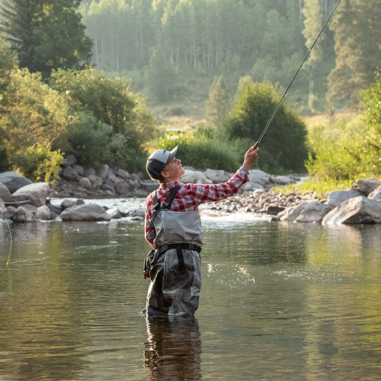 A boy casts a line fly fishing on the calm waters of the Gore Creek in Vail, Colorado. Greenery surrounds them on both sides of the creek.