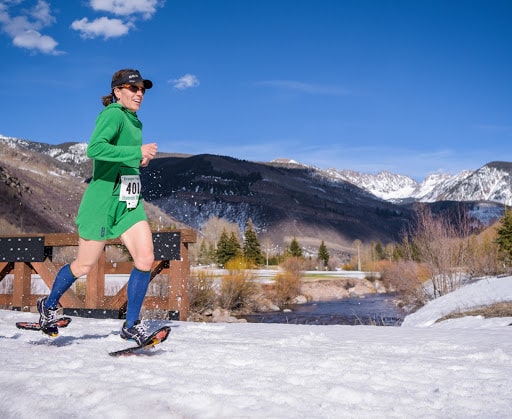 A woman in green running in the snow with mountains in the background.