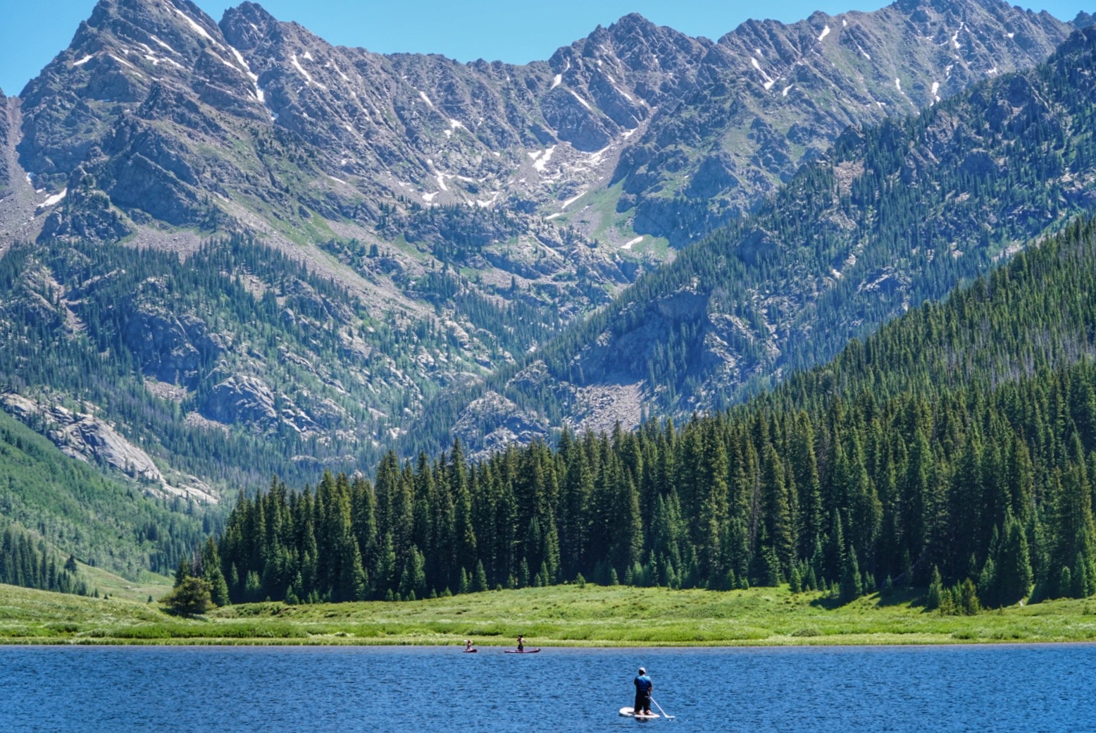 A paddleboarder floats on sapphire Piney Lake. A grassy, tree-covered landscape is behind it and mountains tower above.