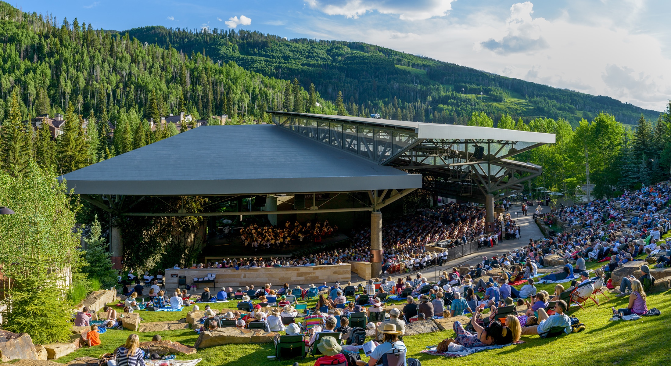 An outdoor concert in Vail at The Amp with mountains in the background. There are many people sitting and laying down on the green grassy field.