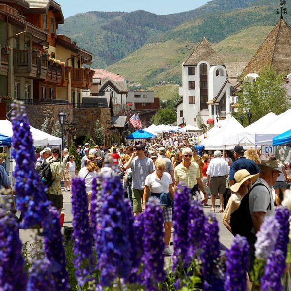 A crowd of visitors and locals stroll through Vail Farmers' Market & Art Show with purple flowers in the foreground and grassy mountainside in the background.