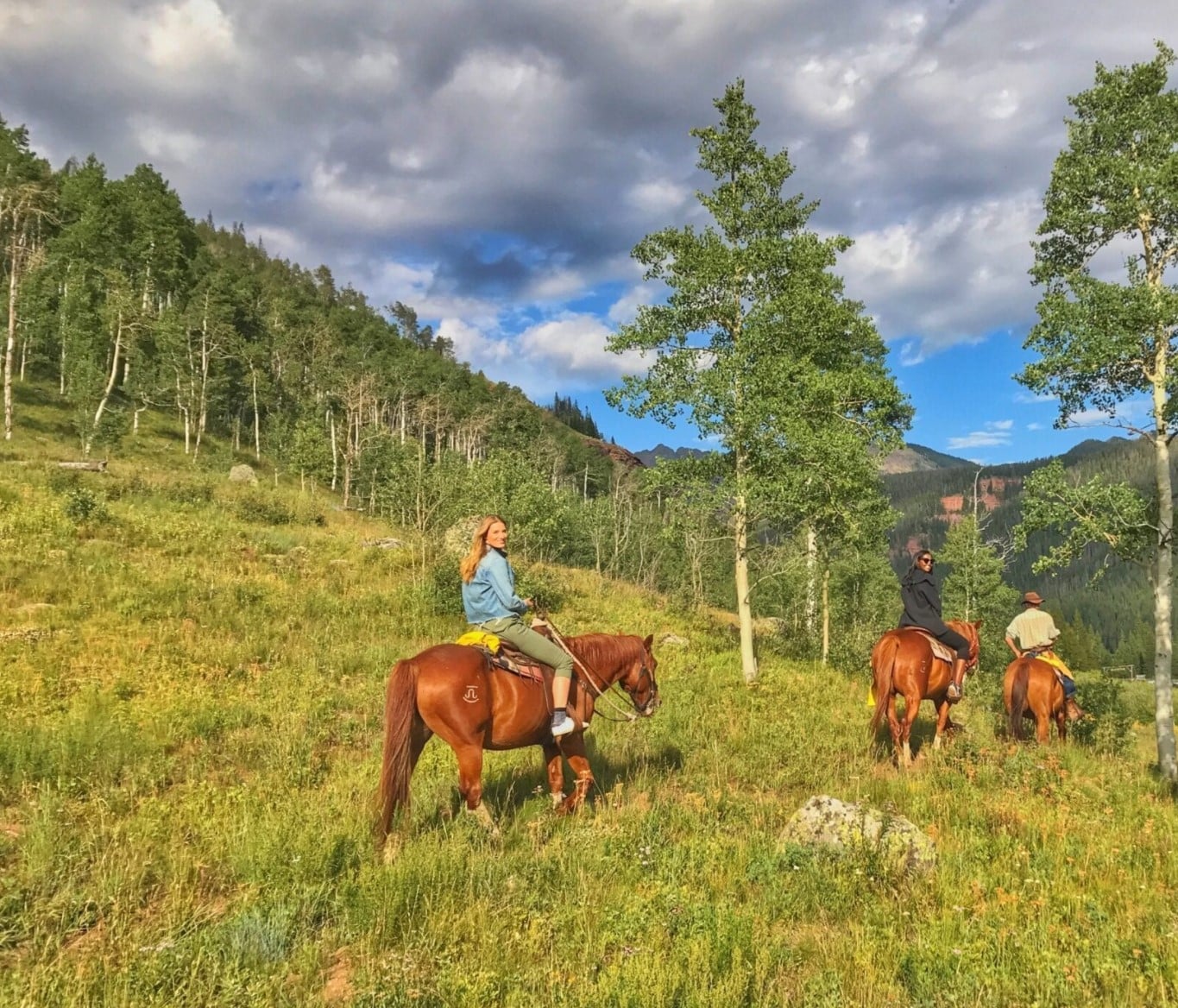 A group of people ride brown horses along a grassy hillside in Vail, Colorado. Trees stand in the background and clouds fill the sky.