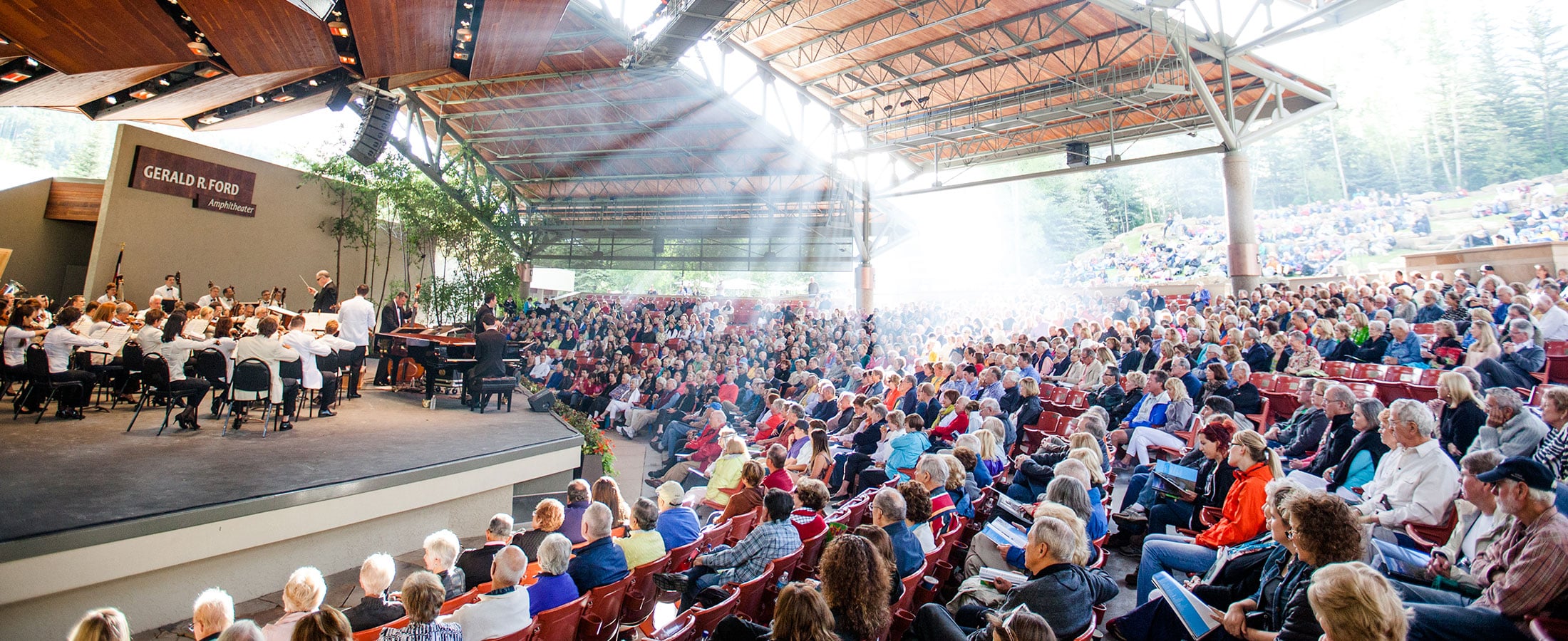 An orchestra performs on the stage of The Amp in Vail. The event attendees are seen sitting quietly and watching the performance, with other attendees sitting farther out on the green grass. Sunlight filters into the open-air amphitheater.