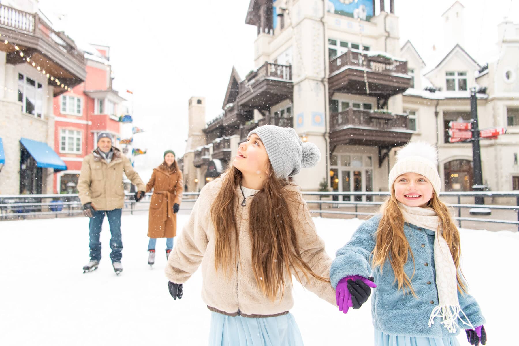A family of four ice skates together in Vail. Two young girls hold hands as they skate in the foreground, and their parents skate in the background.