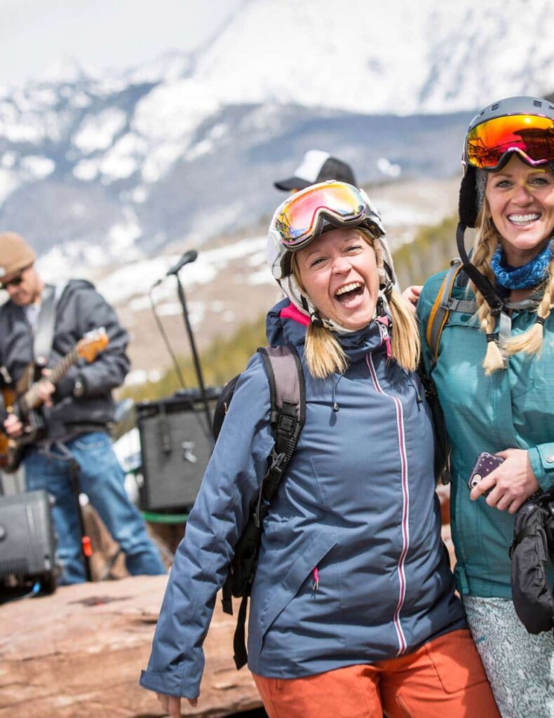 A person wearing a ski helmet and goggles laughs heartily into the camera while hugging a similarly dressed friend while a man with a guitar plays in the background in Vail.