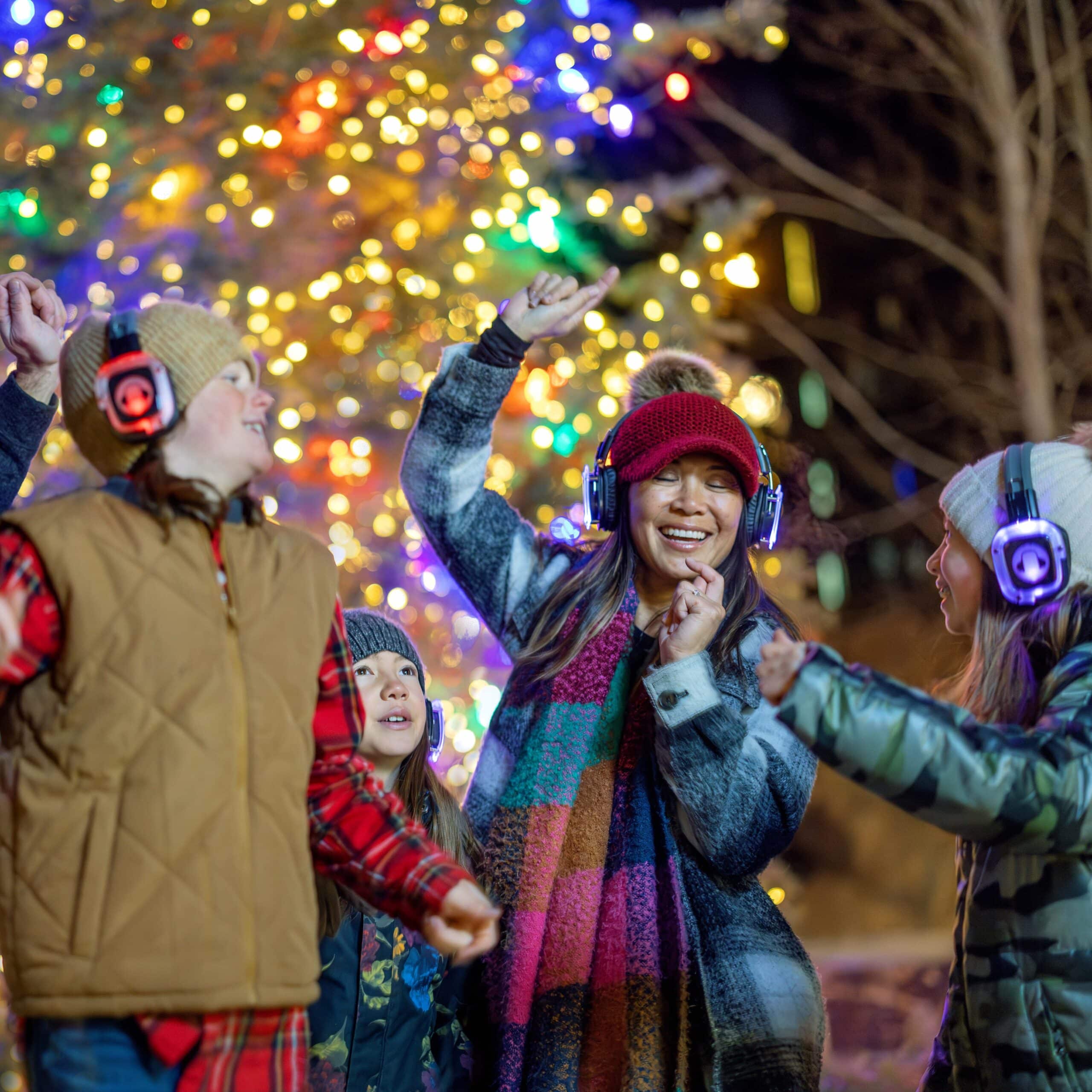 People wearing winter clothes and headphones dance together outdoors at night, with a large, decorated and lit Christmas tree in the background.