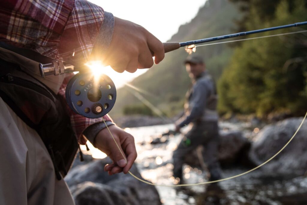 A person wearing a plaid shirt holds a fishing rod in the foreground while someone else fishes in the background. Both are standing in a Vail river with mountains in the background, and the sun is peeking through the fishing pole.