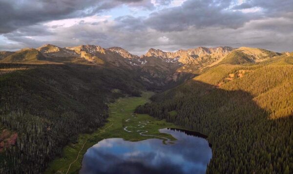 Piney Lake near Vail glimmers in shadows with rugged mountains all around it. The sky is cloudy with just a hint of blue peeking out.