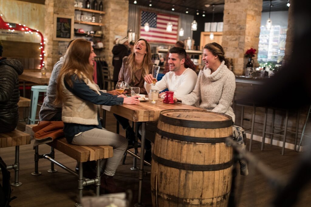 A group of adults is dining at a restaurant. They are laughing and smiling around a table with food and drinks. In the background are fairy lights, an American flag and lantern-looking chandeliers.