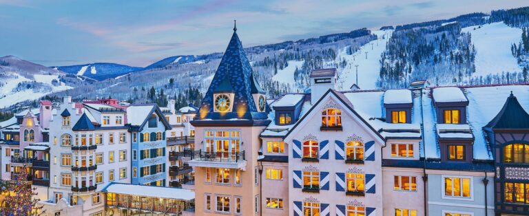 A view of The Arrabelle at Vail Square at dusk, perfect for a Vail vacation rental. Snowy mountain slopes behind the hotel beckon skiers and snowboarders.