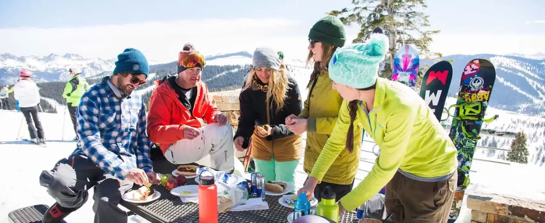 A group of people smile and laugh as they eat lunch by a picnic table on a deck on Vail Mountain. There are snowcapped peaks in the background.