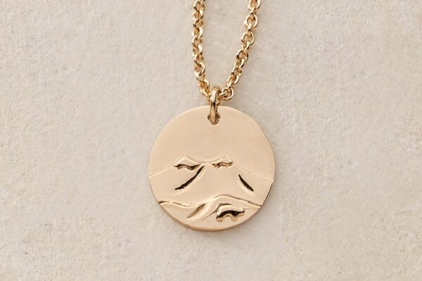 A round yellow-gold pendant featuring a mountain and a bear figure lays on a light-colored background in Vail, Colorado.