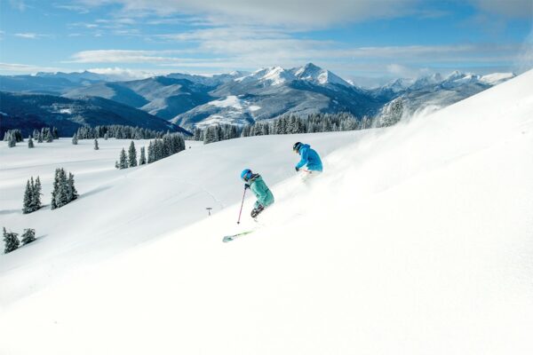 Two skiers in blue jackets schuss down a mountain in Vail. Behind them is a field of snow, a pine forest and majestic mountains.