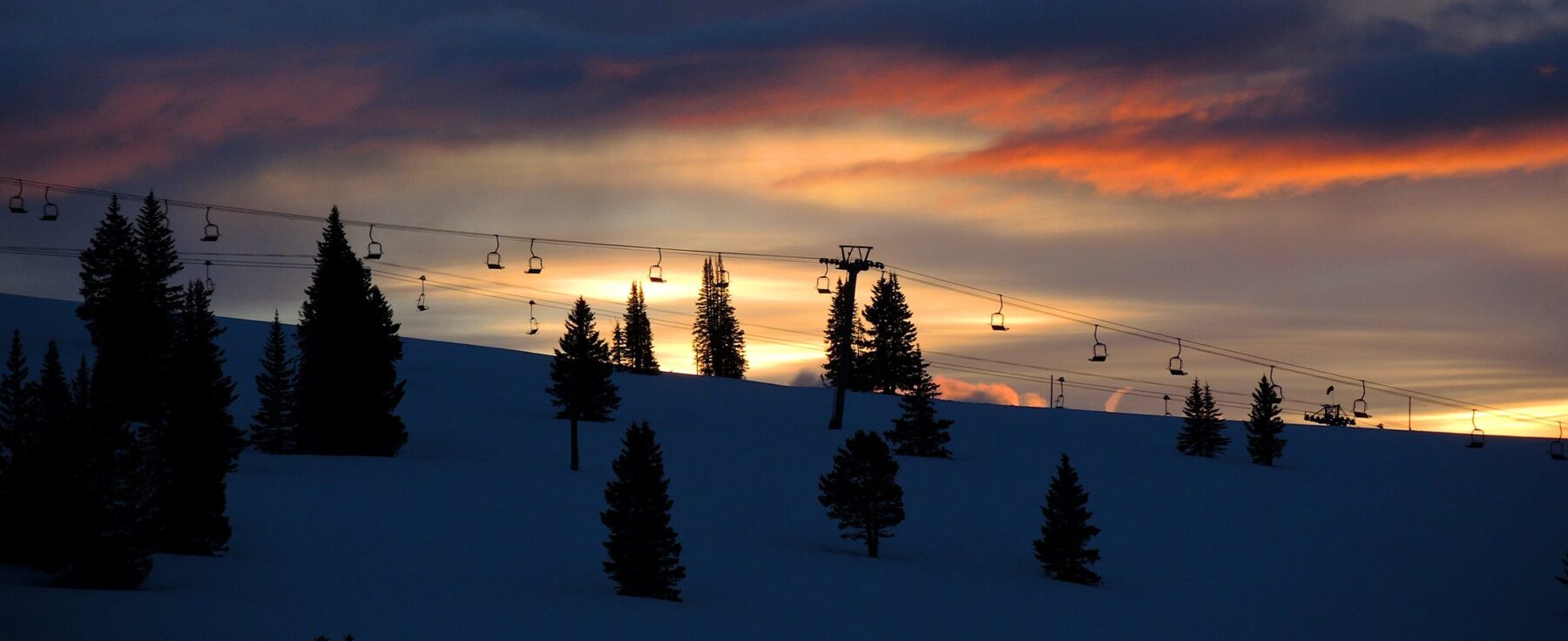 The sun sets on the mountains of Vail, Colorado. Evergreen trees and a ski lift are silhouetted against a sky of yellow, pink and blue.