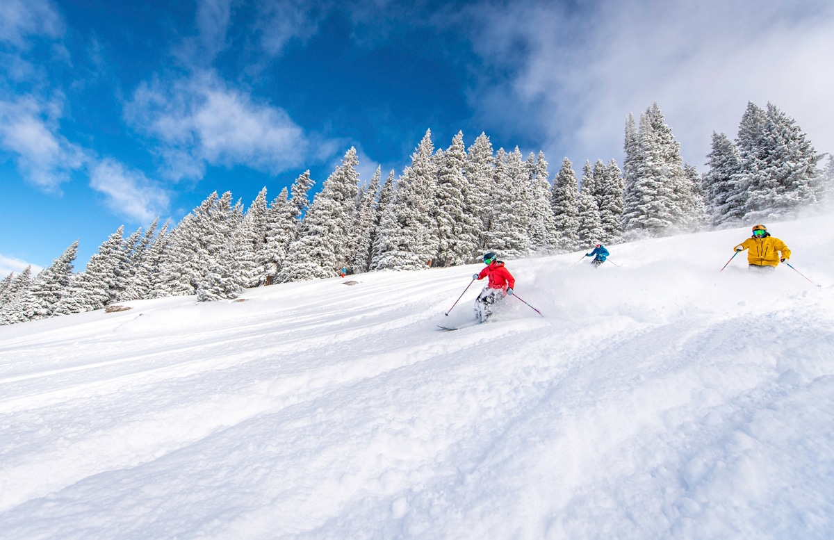 Three skiers in brightly colored winter coats zoom down a snowy Vail Mountain in Colorado. The pine trees behind them are dusted with snow. The sky is mostly blue with whispy clouds.