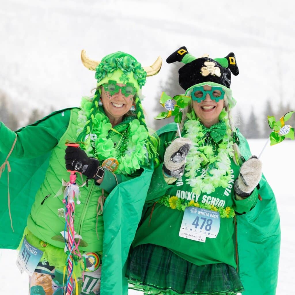 Two people dressed in bright green costumes with hats, glasses, and accessories pose outside in a snowy landscape at a race event.
