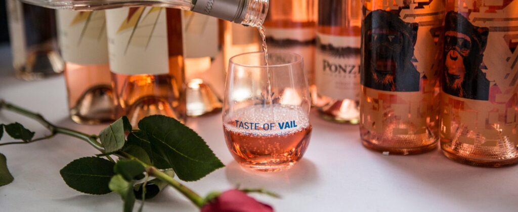 Several bottles of rose wine sit on a table with a white table cloth in Vail, Colorado. Fresh wine is being poured into a stemless wine last marked: Taste of Vail.