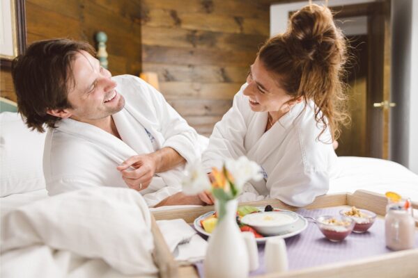 A couple lounges on a Vail Village hotel bed in cozy, white robes. In front of them is a breakfast tray with a plate of assorted fruit and other dishes.