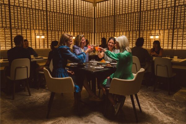 Four friends dressed in semi-fancy outfits sits at a table and clink their cocktails together in a toast at a restaurant in Vail Village.