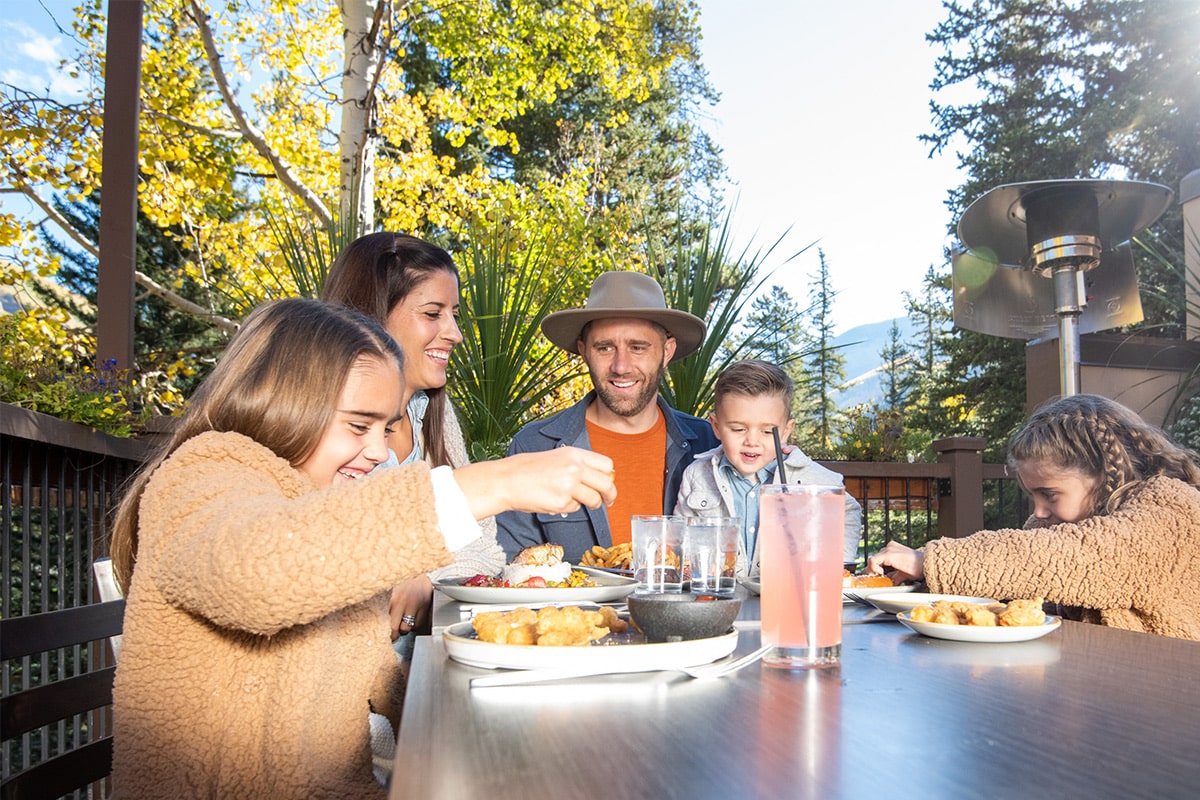 A family of five sits outside beneath autumn-colored trees and share a meal together at an outside patio in Vail Village.