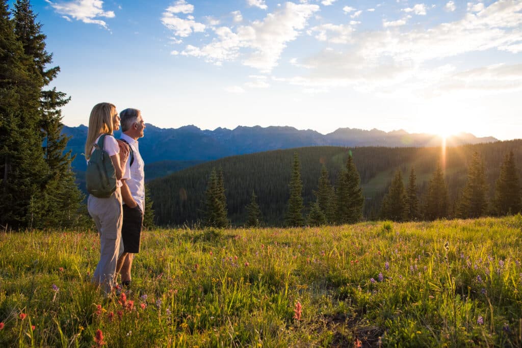 Couple gazing at a mountain range on a hike in Vail, the grass is bright green and speckled with wildflowers.