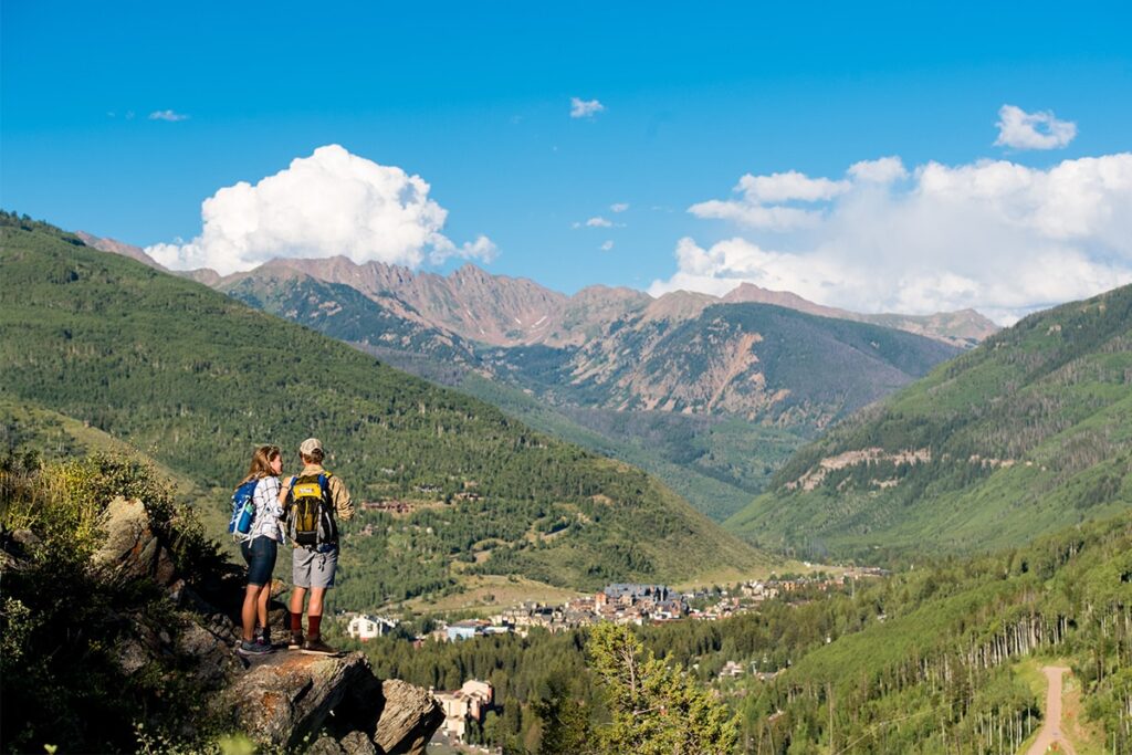 Two people stand on the edge of a rocky outcrop looking down at Vail, Colorado. They are surrounded by lush, green mountain and blue sky.