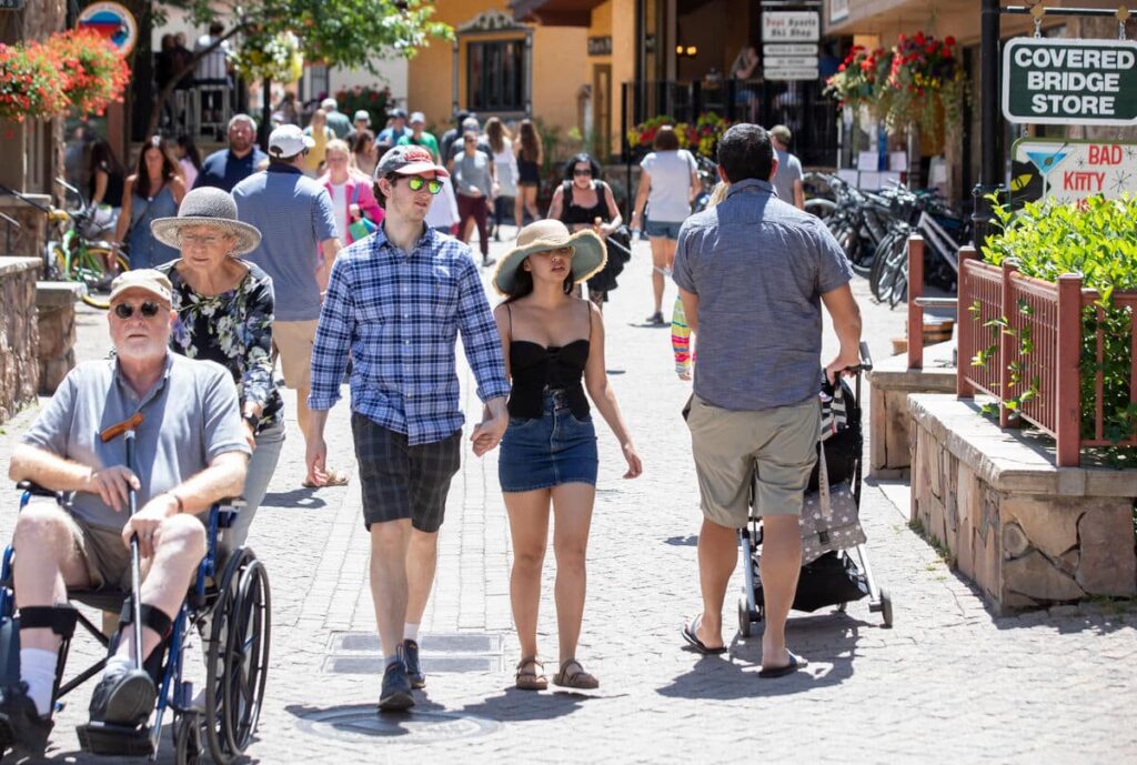 A person in a wheelchair is pushed through Vail Village surrounded by other people on a warm summer day