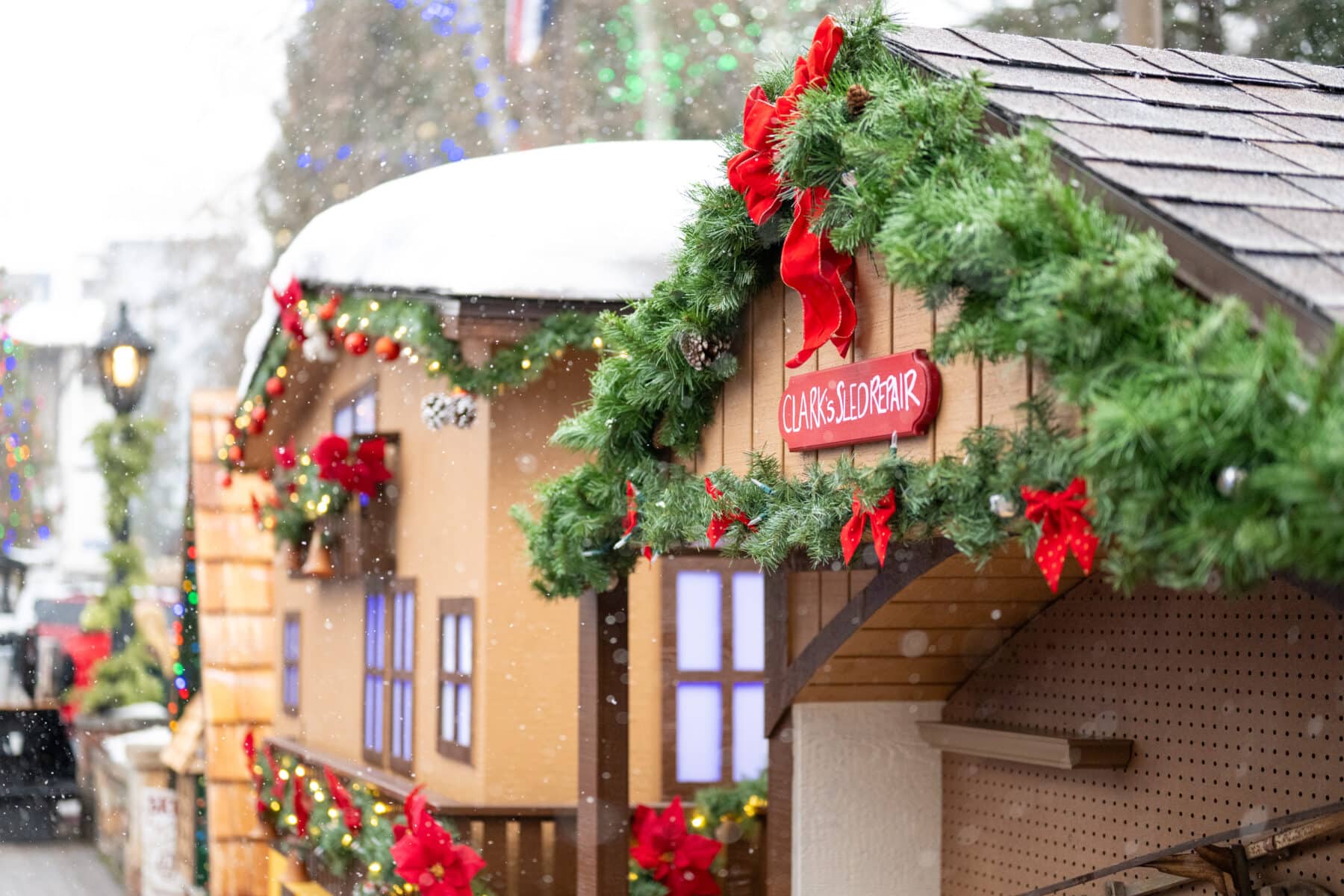 A festive outdoor market display with wooden stalls decorated with green garlands, red ribbons, and poinsettias, with snow on the roofs.