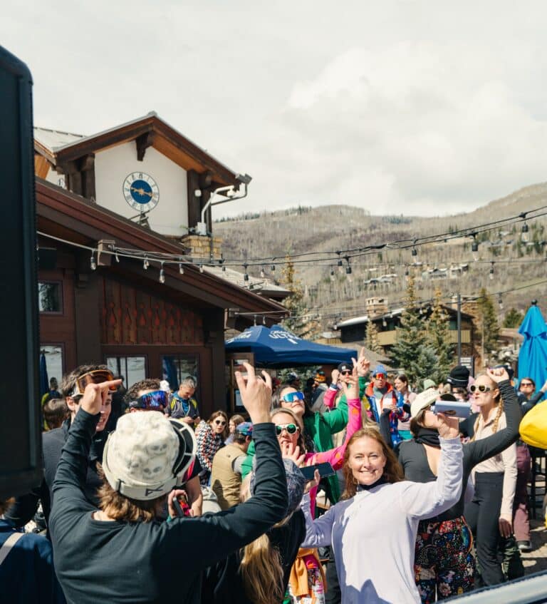 A crowd of people in winter clothing gather outdoors near a wooden building with a clock, raising their arms and enjoying a sunny day.