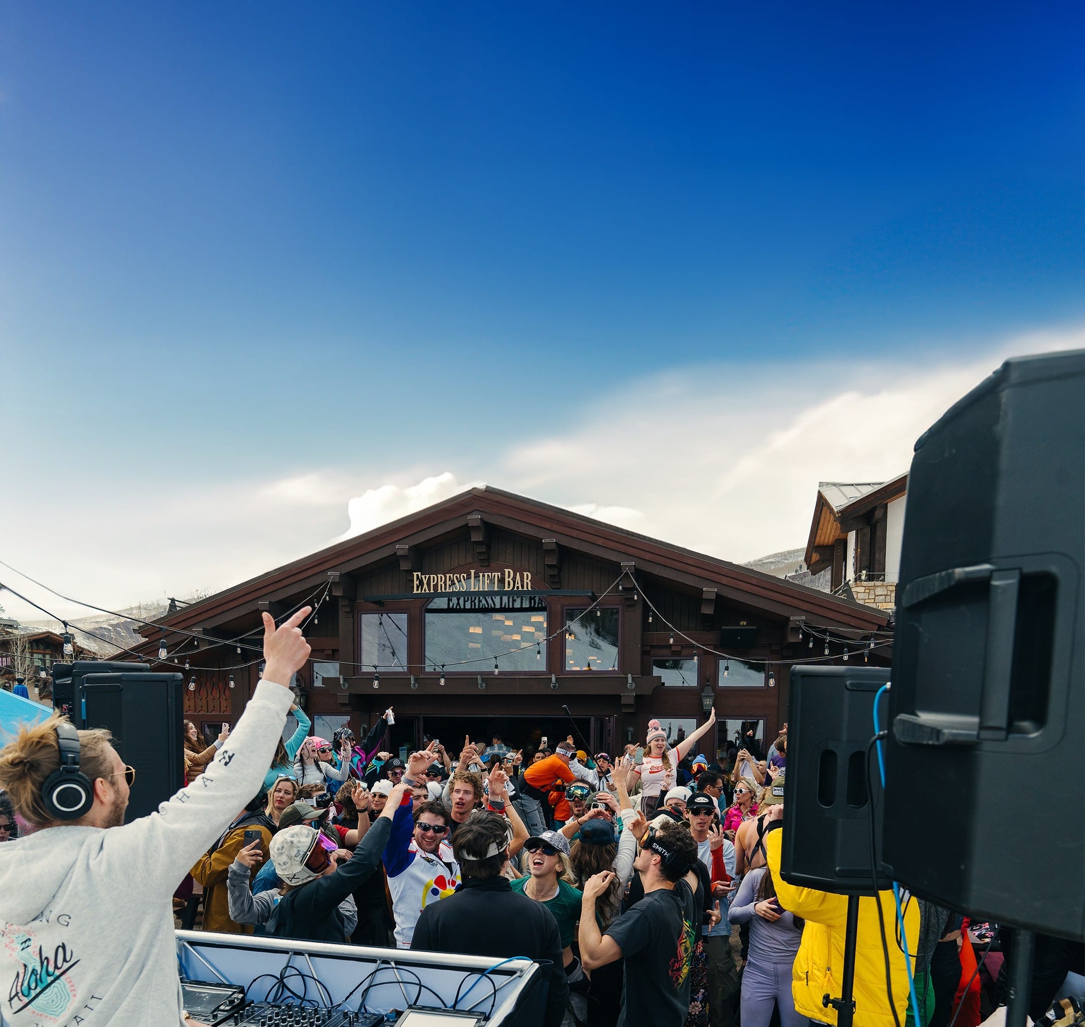 A DJ performs on an outdoor stage in front of a lively crowd at Express Lift Bar, with clear blue sky overhead and wooden buildings in the background.