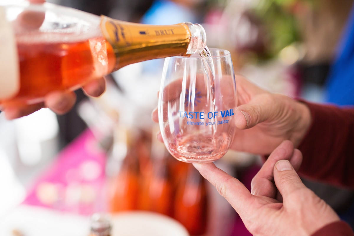 A person pours a blush wine into a stemless wine glass that says, "Taste of Vail."