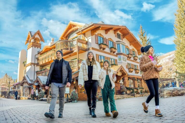 Two parents and their two children walk through the sunny cobblestone streets of Vail, Colorado, with Tyrolean-style buildings in the background.