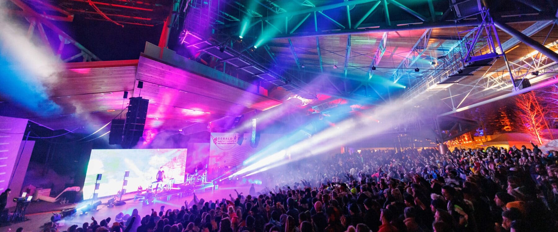 Colorful lights and smoke illuminate the ceiling of the Gerald R. Ford Amphitheater while two drummers stand on stand before a large ground.