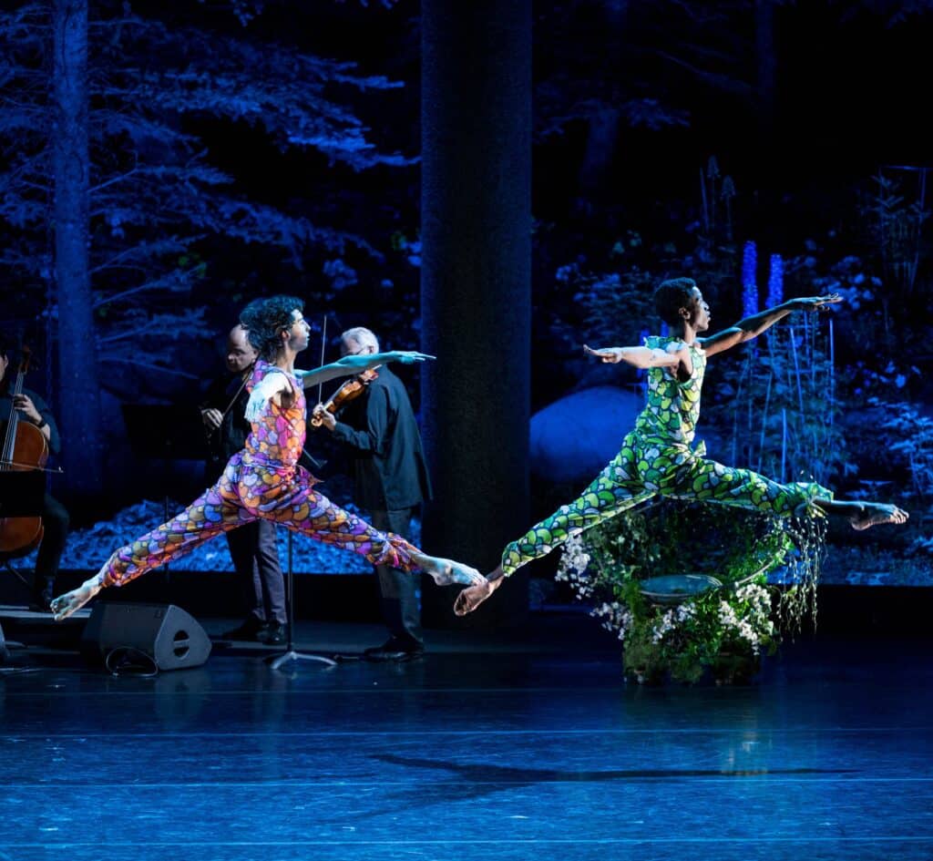 Two ballet dancers in colorful costumes leap across a stage, accompanied by live string musicians, with a forest-themed backdrop behind them.