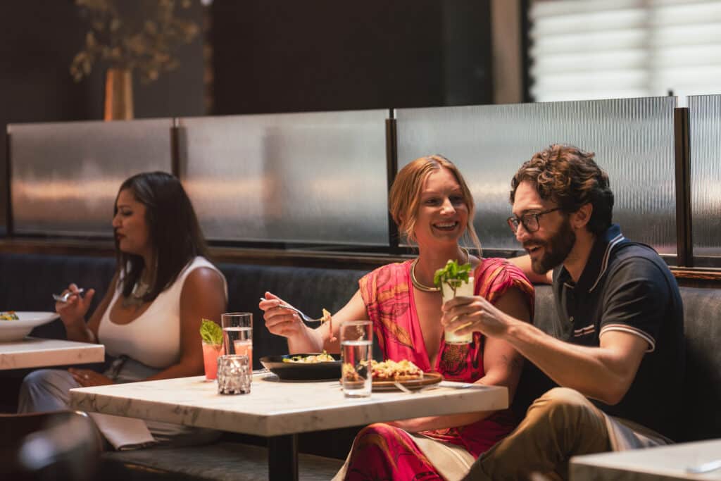 A young couple sit at an upscale restaurant. The man is holding a drink with a garnish and the woman is holding a fork. She is looking at him with a smile while he talks.