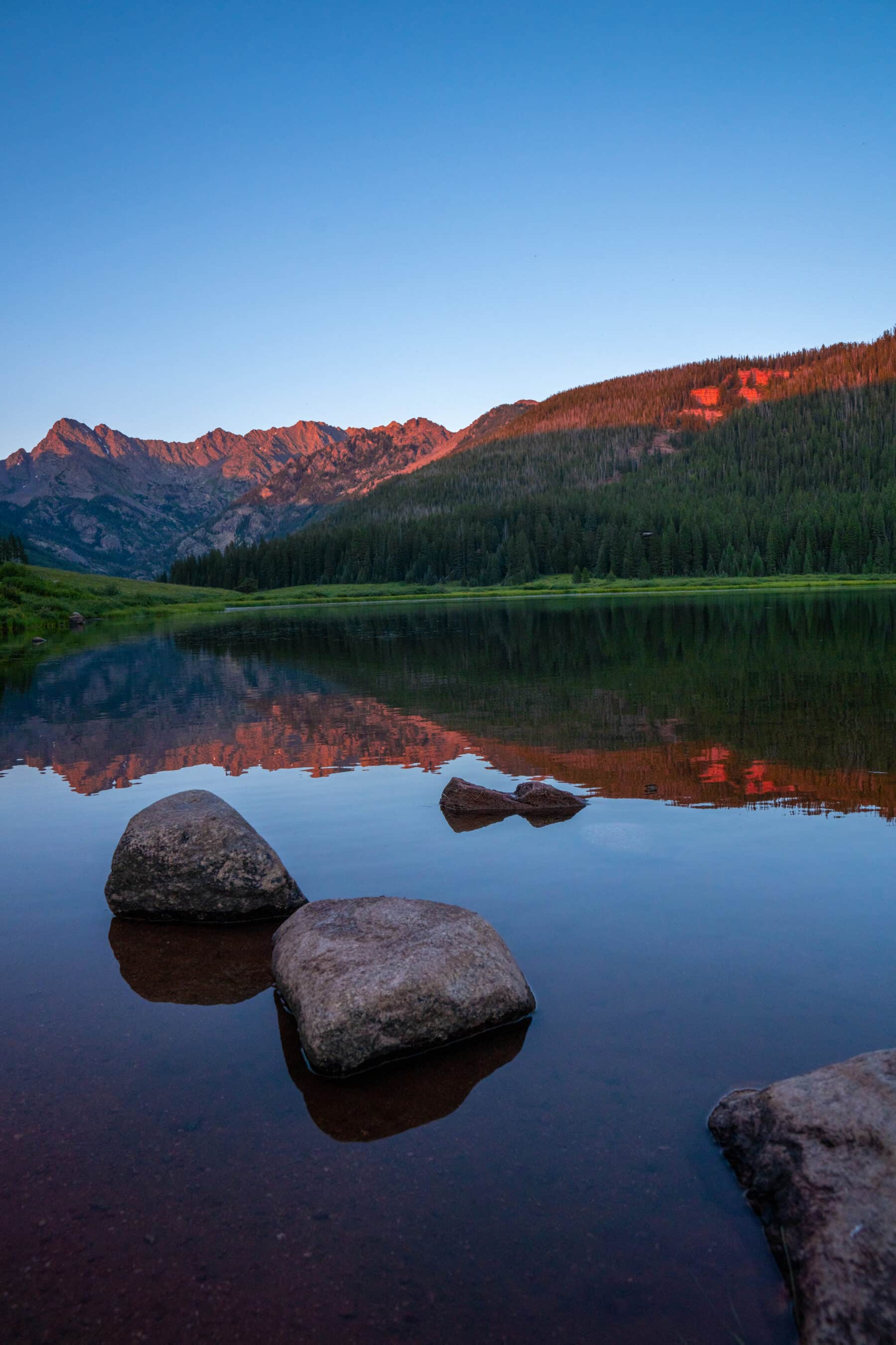 Light shining over Piney Lake. The tops of the far mountains glow bright red. There is lush grass on the banks of the lake while the close up of the water below reflects the landscape.