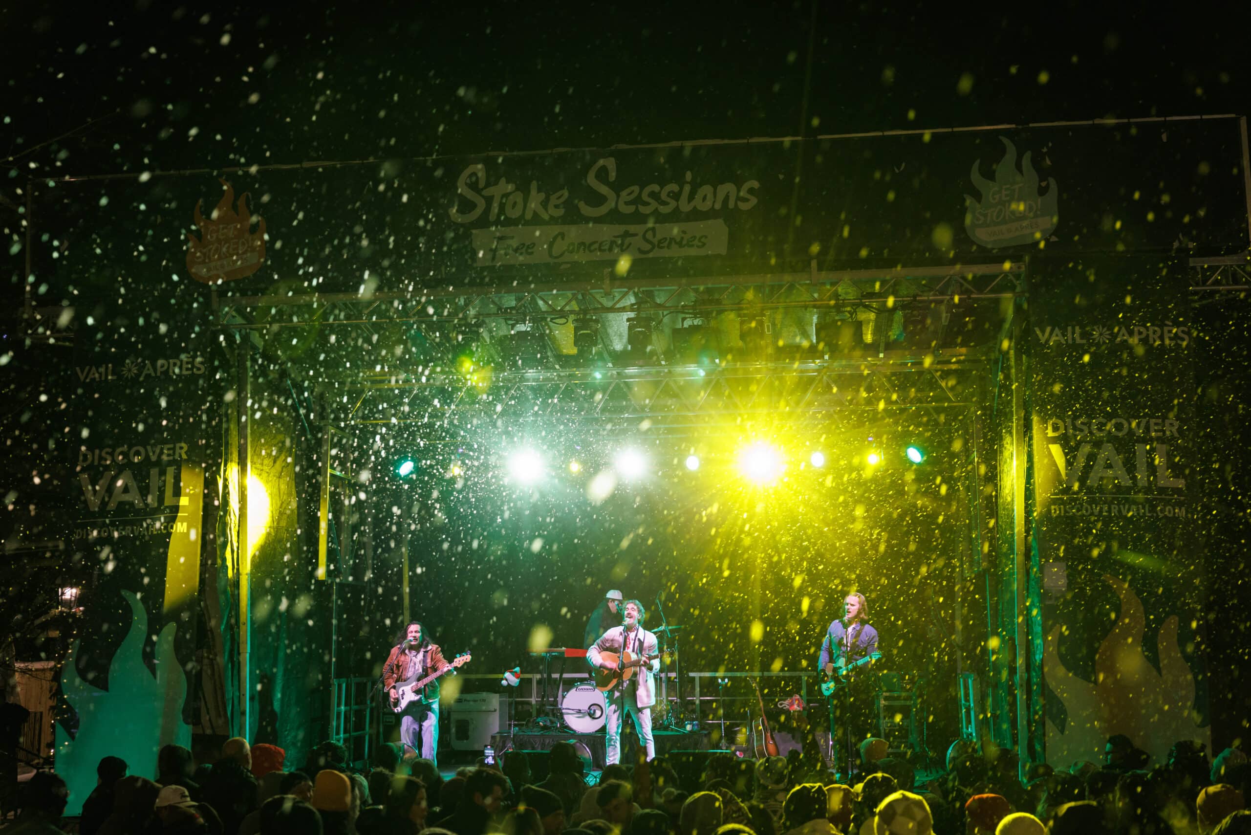 A band performs on an outdoor stage at night under bright lights, with artificial snow falling and a crowd gathered in front.
