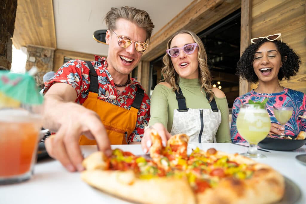 Two people reach for slices of freshly-baked pizza as they eat and chat with friends in Vail, Colorado.