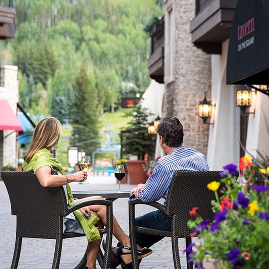 View from behind of a man and woman sitting at an outdoor table at the Tavern on the Square in Lionshead Village, drinking wine and looking to the mountains in Vail, Colorado