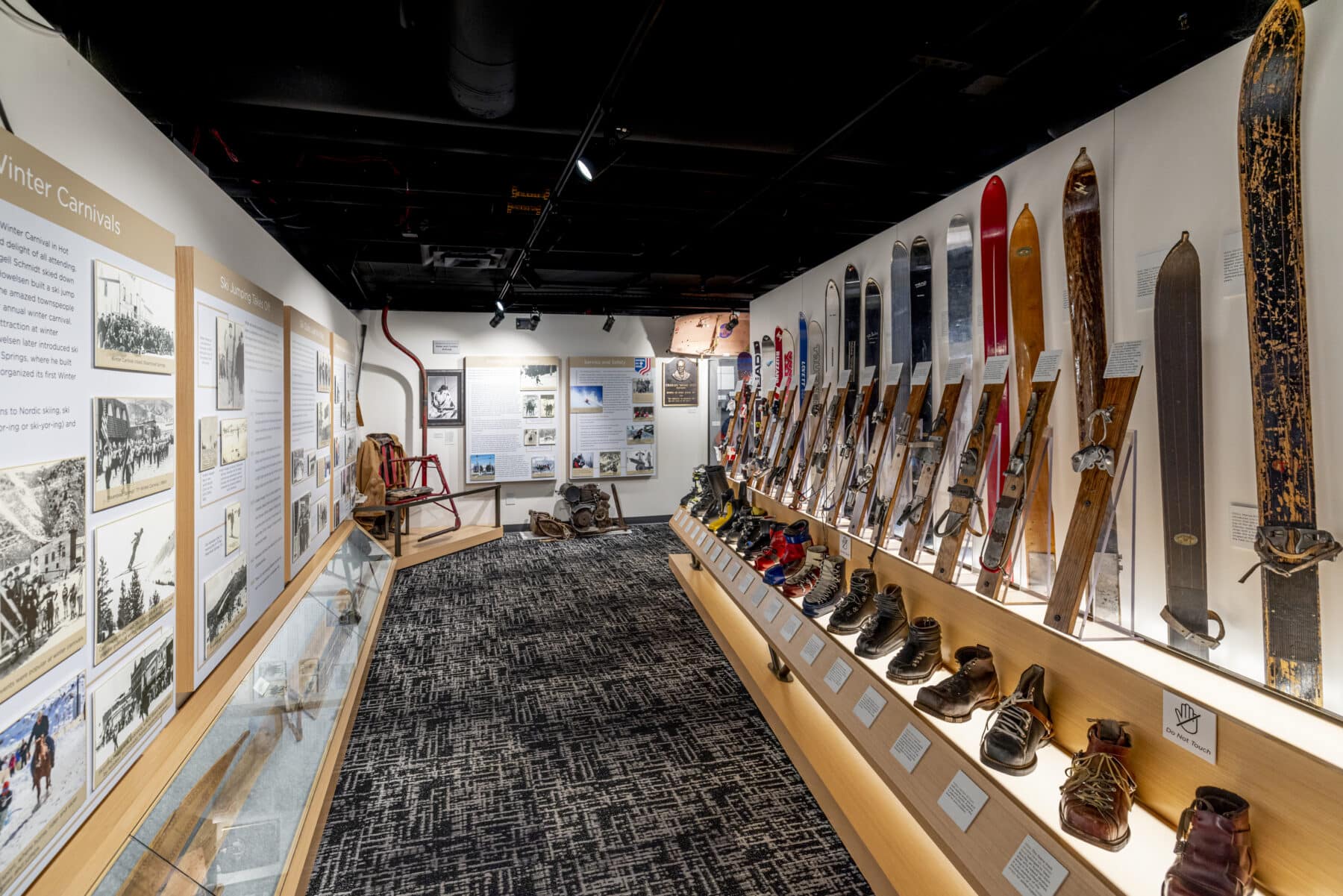 A museum exhibit displays a variety of vintage skis, boots, and historical winter sports equipment, with informational panels and photographs on the walls.