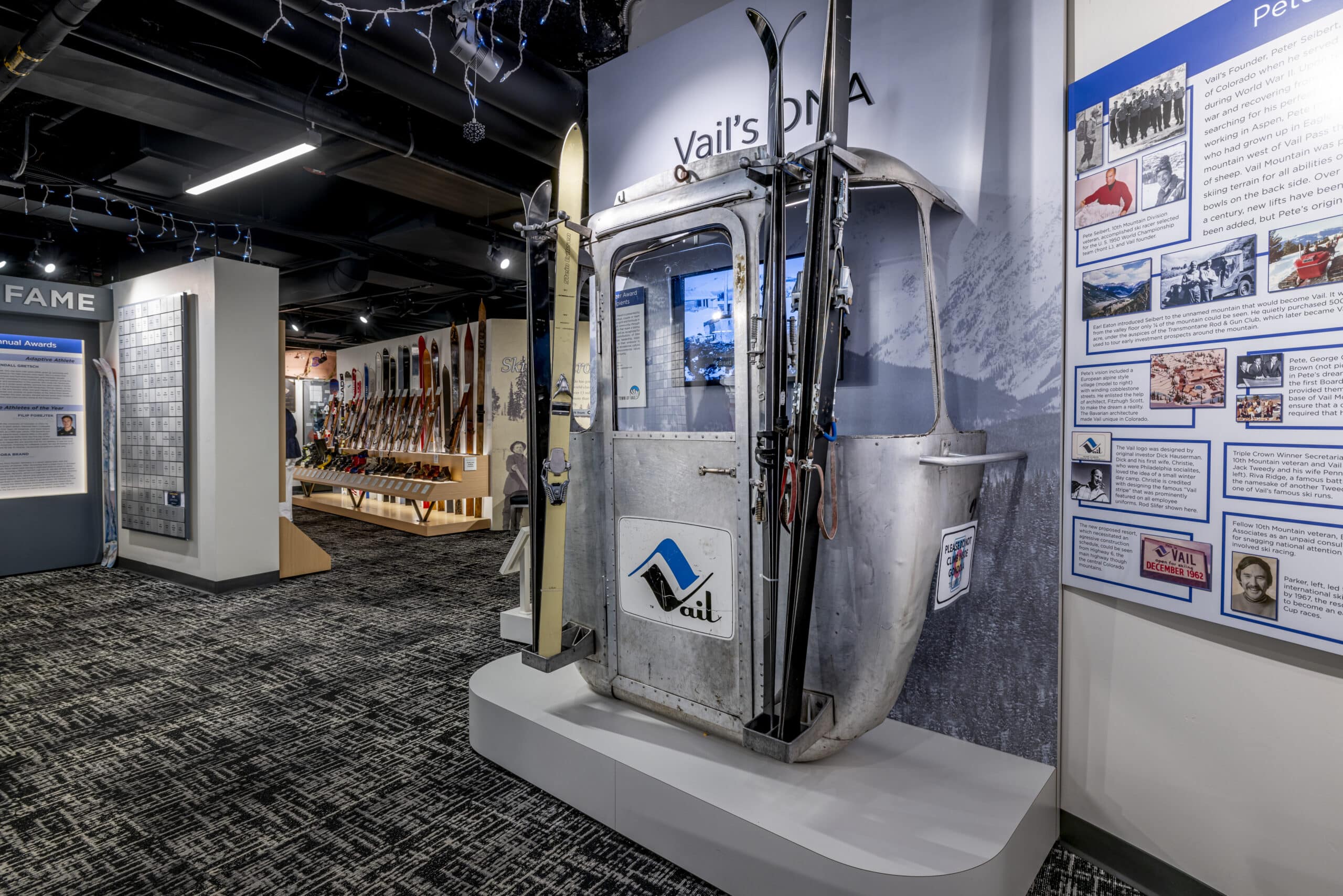 A vintage ski gondola is displayed in a museum exhibit, surrounded by informational panels, historical photos, and ski-related items.