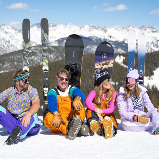A group of four friends wearing colorful ski gear bask in the sun, laughing together, at Vail, Colorado. The sky is blue and the mountains are in the background.