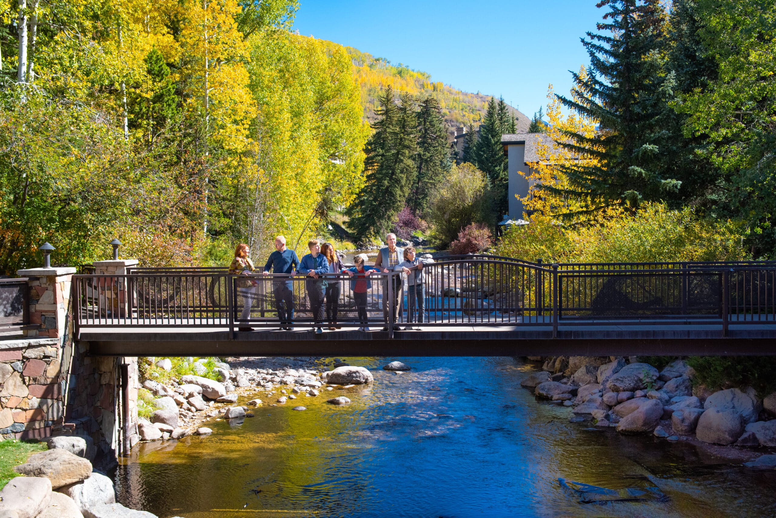 A family of seven stands on a bridge in Vail that spans Gore Creek. Behind them, yellow and green aspen trees rise up and are reflected in the creek's waters.